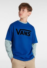 Sininen puuvillapaita, jossa on suuri musta "VANS" logo. Paita on puettuna vaaleansinisen pitkähihaisen paidan päälle ja se on yhdistetty beigeihin housuihin.