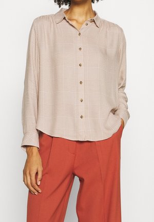 Femme portant une chemise à manches longues boutonnée à carreaux beige, rentrée partiellement dans un pantalon taille haute de couleur rouille, main dans la poche.