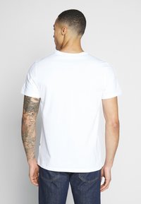 Tricou alb din bumbac, cu mâneci scurte, guler rotund și design simplu. Prezintă tatuaje vizibile pe braț și blugi albaștri.
