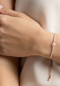 Delikat armband i roséguld med en hjärtformad, rosa sten i mitten, omgiven av klara kristaller och en tunn kedja med hängande detaljer.