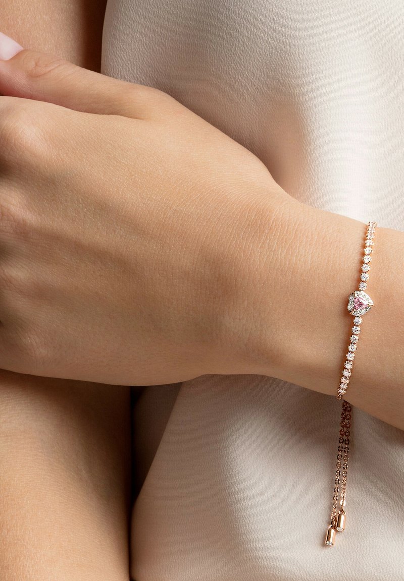 Delikat armband i roséguld med en hjärtformad, rosa sten i mitten, omgiven av klara kristaller och en tunn kedja med hängande detaljer.