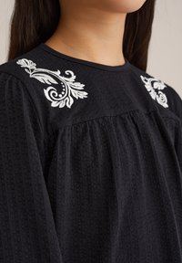 Mustmust värviline pluus, millel on tekstuurne kude, puffseelikud ja valged lillepärlite tikandid õlgadel. Varruka ümber on pleeditud disain.