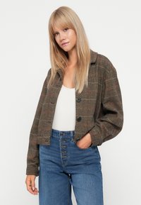 Veste courte à carreaux bruns avec un col, deux boutons noirs et une coupe décontractée, associée à un jean bleu taille haute. Texture anti-bouloche.