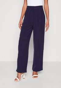 Marineblauwe wijde broek met een hoge taille, voorklappen en een gladde stoftextuur, gecombineerd met lichtgekleurde sandalen met hak.