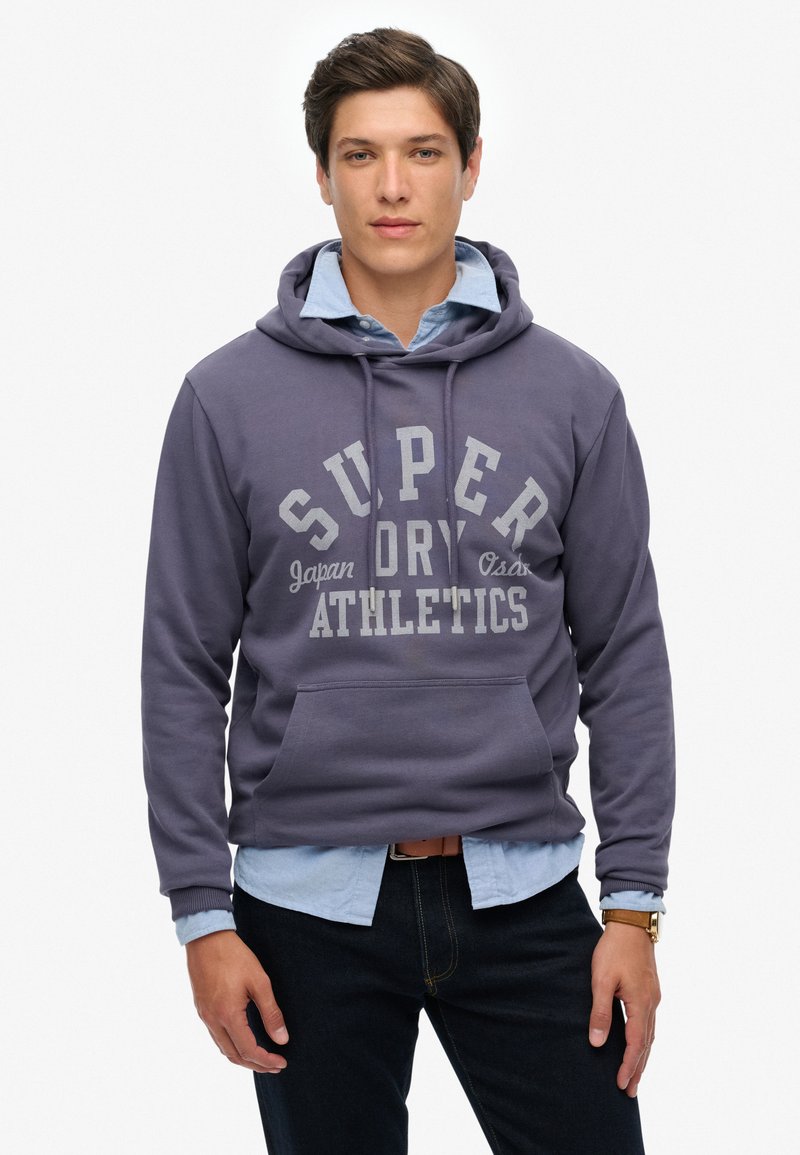 Sudadera morada con el texto "SUPER DRY ATHLETICS", material de algodón, bolsillo tipo canguro, capucha con cordón, llevada sobre una camisa de color azul claro.