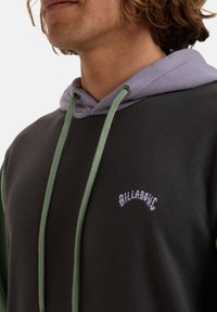 Sudadera con capucha de color verde oscuro, capucha morada y cordones verdes. Presenta el logotipo bordado "BILLABONG" en púrpura claro.