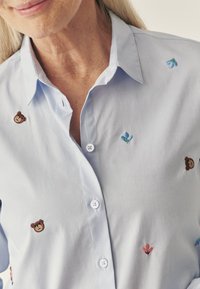 Jasnoniebieska koszula z guzikami z haftowanym wzorem misia i kwiatów. Miękka tekstura, klasyczny kołnierzyk oraz widoczna listwa guzikowa.