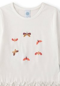 Camicia bianca a maniche lunghe con orlo a volant. Presenta sei stampe di farfalle colorate in arancione, giallo e marrone, centrato sul davanti.