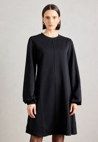 Zwarte jurk met lange mouwen, een ontspannen pasvorm, ronde halslijn en gathered manchetten. Soepele textuur met een detail van een centrumnaad.