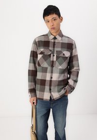 Brun og grå rutete flanellskjorte med knappelukking foran, to brystlommer og krage, kombinert med blå jeans og en beige skulderveske.