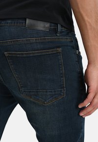 Mörkblå denimjeans med en läderlapp vid midjan, rak passform, och två bakfickor med kontrasterande sömmar.