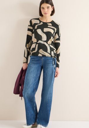 Top nero a maniche lunghe con motivo astratto beige, abbinato a jeans blu a gamba larga. Scarpe da ginnastica bianche e giacca color bordeaux in mano.