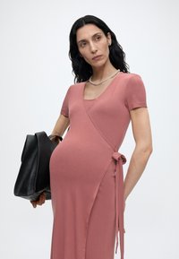 Gravid kvinne med langt mørkt hår har på seg en støvete rosa omslagskjole og perlekjede, og holder en stor svart skinnveske.