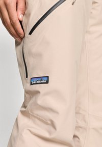 Μπεζ παντελόνι με λεία, ματ ύφανση, τσέπες με φερμουάρ και μαύρη επένδυση, καθώς και ετικέτα λογότυπου Patagonia στο μηρό.