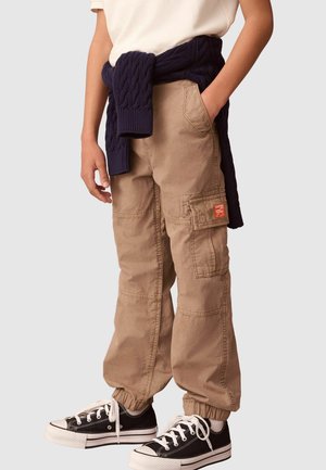 Pantaloni cargo khaki con polsini elasticizzati e molteplici tasche, abbinati a un maglione lavorato a maglia blu scuro legato in vita e scarpe da ginnastica nere.