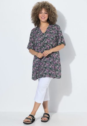 Smilende kvinde med krøllet hår iført en mønstret tunika, hvide korte bukser og sorte sandaler, stående mod en ensfarvet hvid baggrund.