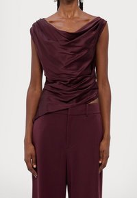 Top drapé bordeaux en tissu lisse, avec un col en cowl et des manches courtes, associé à un pantalon fuselé assorti.