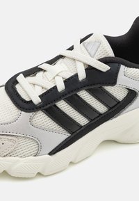 Zapatilla deportiva en blanco y negro con parte superior de malla, suela de goma y un prominente diseño de tres rayas en negro. Incluye detalles en plateado.