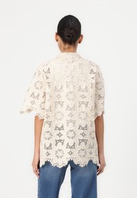 Blusa in pizzo color avorio con motivo floreale, maniche corte e orlo a merletto. Tessuto trasparente, design con colletto e vestibilità ampia.