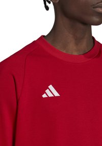 Rode sweatshirt met een ronde hals, voorzien van geribbelde accenten en een wit Adidas-logo op de bovenste linkerzijde. Glad textuur.
