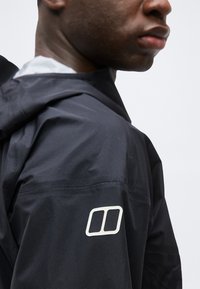 Gros plan d'une personne portant une veste noire avec un logo double rectangle blanc sur le haut du bras.
