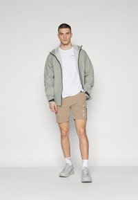 Ljust grönt vattentätt jacka, vit t-shirt, beige shorts med brodyr och grå sneakers. Enkel design, sportig stil.