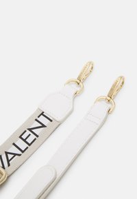 Correia branca com acessórios em metal dourado, um corpo em tela e uma seção em tecido bege com a palavra "VALENTINO" em letras pretas em negrito.