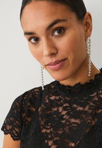 Sølvøredobber med vedhæng i et kaskadedesign, med klare stenaccenter; kombineret med en sort blondebluse med blomsterprint.