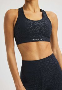 Atletische persoon in een zwarte, gebloemde sport-bh en hoge taille leggings, die een getonede buik laat zien, staande voor een effen achtergrond.