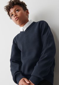 Tmavomodrý sveter s výstrihom do "V", vyrobený z mäkkého materiálu. Má rebrované manžety a lem, nosený cez bielu košeľu s golierom.