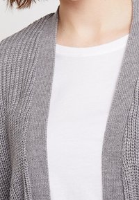 Szary dziany kardigan z teksturowanym wzorem, z otwartym przodem i prążkowanymi krawędziami, noszony na zwykłej białej koszulce.
