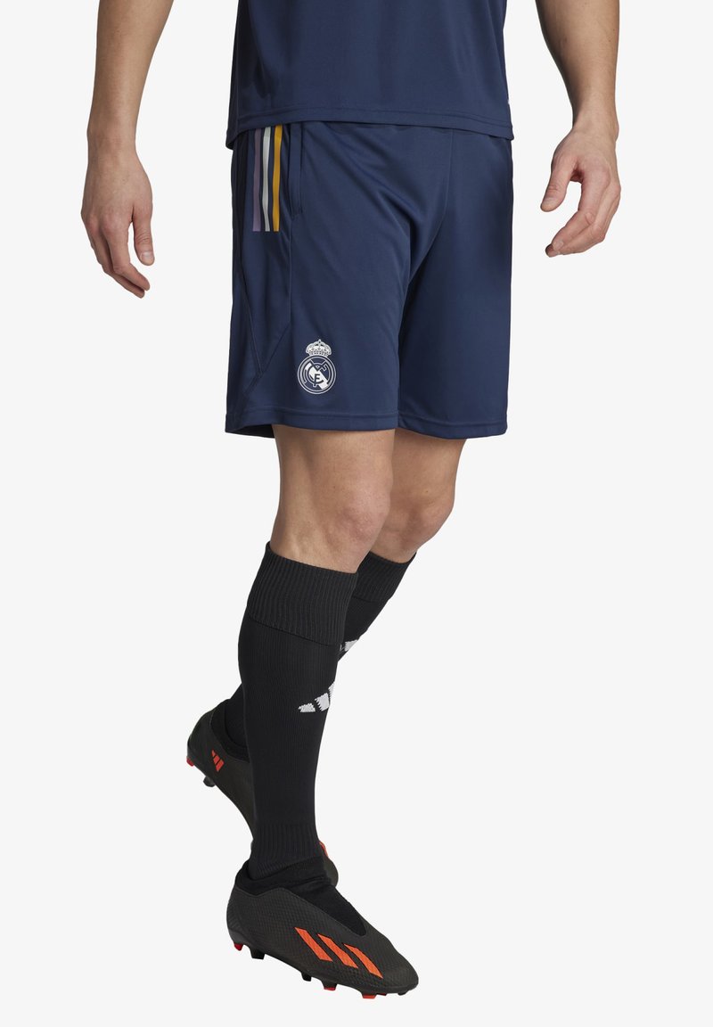 Pantalones cortos de fútbol de color azul marino con rayas laterales amarillas, moradas y blancas, y el logo del Real Madrid. Se llevan con calcetas negras hasta la rodilla y botas negras.