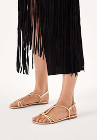 Szalagos, bézs lapos talpú szandálok, T-pántos kialakítással és aranyszínű díszítésekkel. A háttérben fekete rojtos anyag látható.