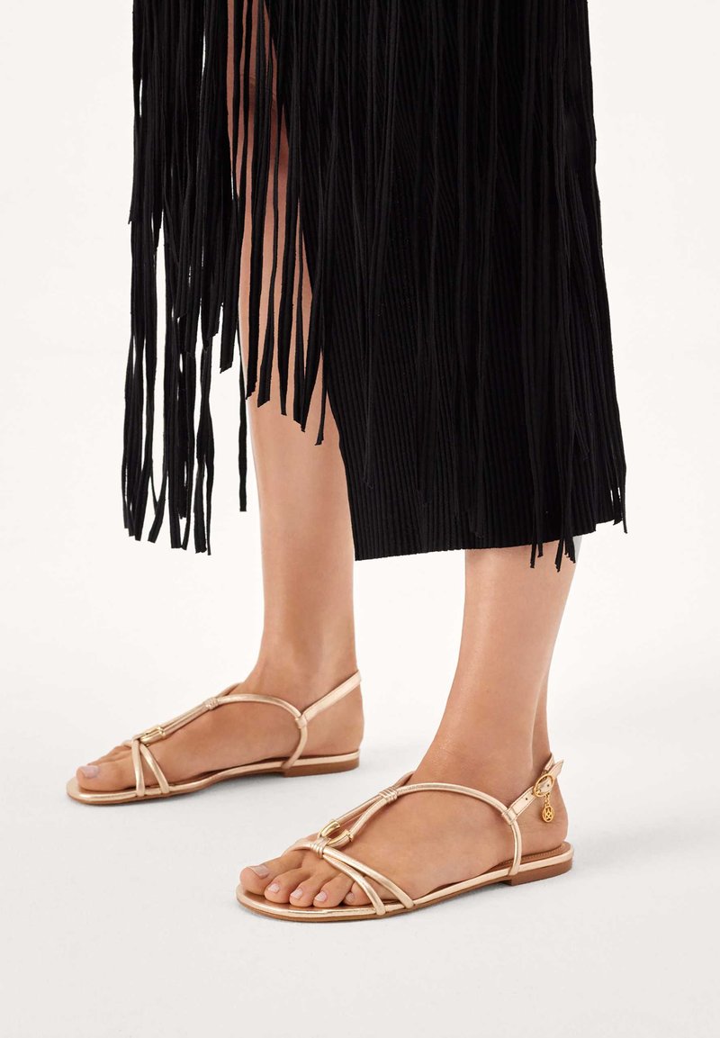 Szalagos, bézs lapos talpú szandálok, T-pántos kialakítással és aranyszínű díszítésekkel. A háttérben fekete rojtos anyag látható.