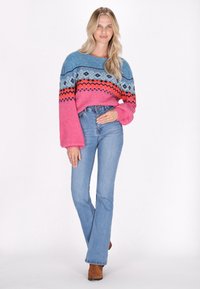 Mehrfarbiger Strickpullover in Blau, Pink und Orange mit geometrischen Mustern, kombiniert mit hellblauen Schlagjeans und braunen Stiefeletten.