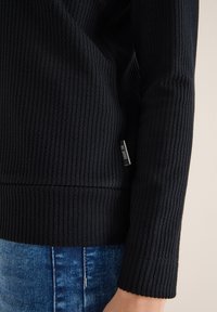 Maglione nero in tessuto a coste con design testurizzato e un'etichetta con la scritta "GAME ON", abbinato a jeans di denim. Le estremità delle maniche sono leggermente svasate.