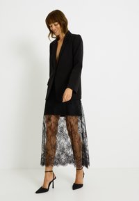 Blazer noir porté sur une robe à volants en dentelle. La robe présente une superposition en dentelle transparente avec des motifs floraux, associée à des escarpins noirs à bout pointu.