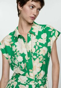 Groene bloemenjurk met korte mouwen, kraag en knoopsluiting aan de voorkant. Witte bloemen met beige accenten op een gestructureerde stof.