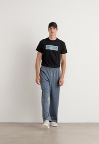 Camiseta negra con texto azul, pantalones de chándal grises y zapatillas blancas, complementados con una gorra negra. Tela suave y diseño de corte relajado.