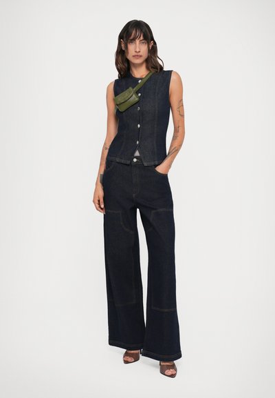 Top sem mangas de denim escuro com frente de botões, combinado com calças largas de denim escuro. Apresenta uma bolsa transversal verde e sapatos de ponta aberta.