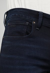 Mörkblå denimjeans med synliga bältesöglor, nitar och en liten framficka, bärs med en vit skjorta.