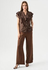 Blouse brune à volants avec des boutons et un pantalon large brun. Texture douce et design fluide, complété par des talons fins noirs.