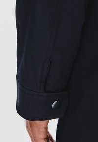 Mankiet granatowej kurtki o gładkiej teksturze, z detalem z zatrzaskiem i widocznym szyciem wzdłuż szwu.