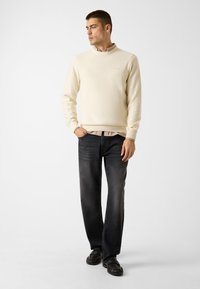 Maglione crema chiaro con collo arrotondato, abbinato a jeans grigio scuro. Il modello indossa scarpe nere, in piedi davanti a uno sfondo neutro.
