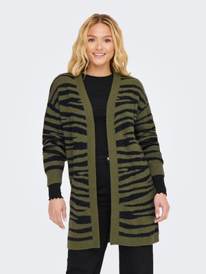 Femme portant un long cardigan rayé vert et noir ouvert devant, sur un top noir et un pantalon noir, souriante avec les mains détendues sur les côtés.