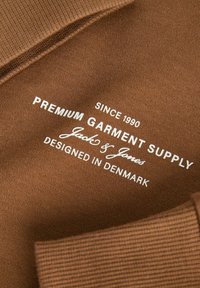 Brown fabric label featuring white text. It reads "PREMIUM GARMENT SUPPLY," "Jack & Jones," and "DESIGNED IN DENMARK," with a ribbed texture.