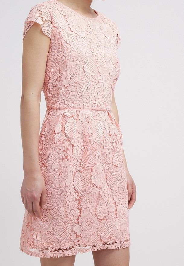 Robe rose longueur genou avec motif en dentelle florale, manches courtes et taille ajustée, portée par une personne sur un fond uni.