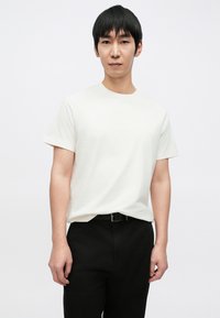 Bílé tričko s kulatým výstřihem a krátkými rukávy z bavlny, kombinované s černými kalhotami. Jednoduchý design, hladká textura a volný střih.