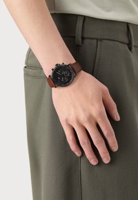 Reloj cronógrafo negro con una esfera redonda, correa de cuero marrón y tres subesferas en una muñeca, combinado con pantalones verdes.