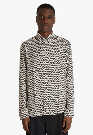 Uomo che indossa una camicia a maniche lunghe con bottoni e motivo a zigzag bianco e nero, in piedi davanti a uno sfondo semplice.